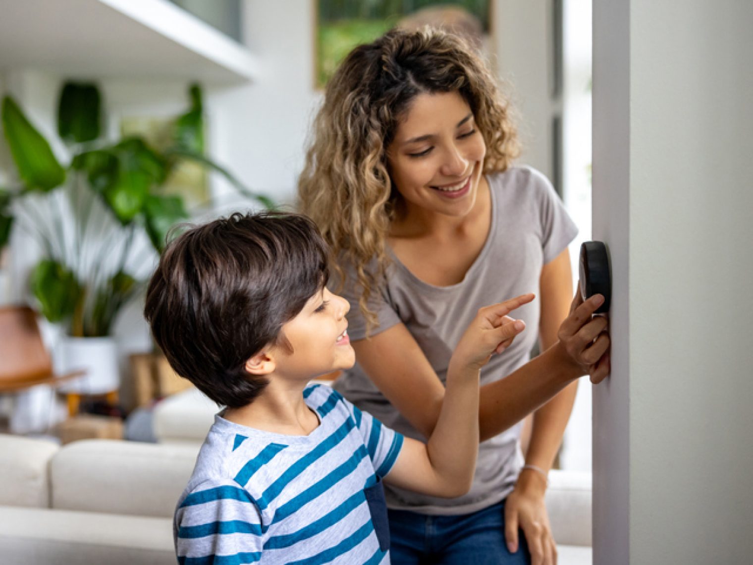 A smiling young boy in a striped blue and white T-shirt looks up at a woman who is pointing to a digital thermostat on the wall.