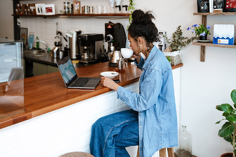 A woman sitting at a coffee bar drinking her cup of coffee and looking at her laptop.