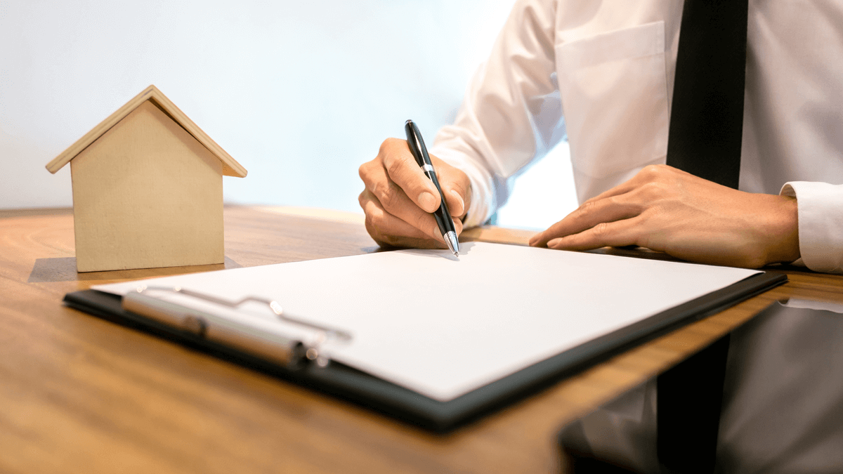 Person in a white shirt and black tie signing a document on a clipboard at a wooden desk with a small house model in the background.